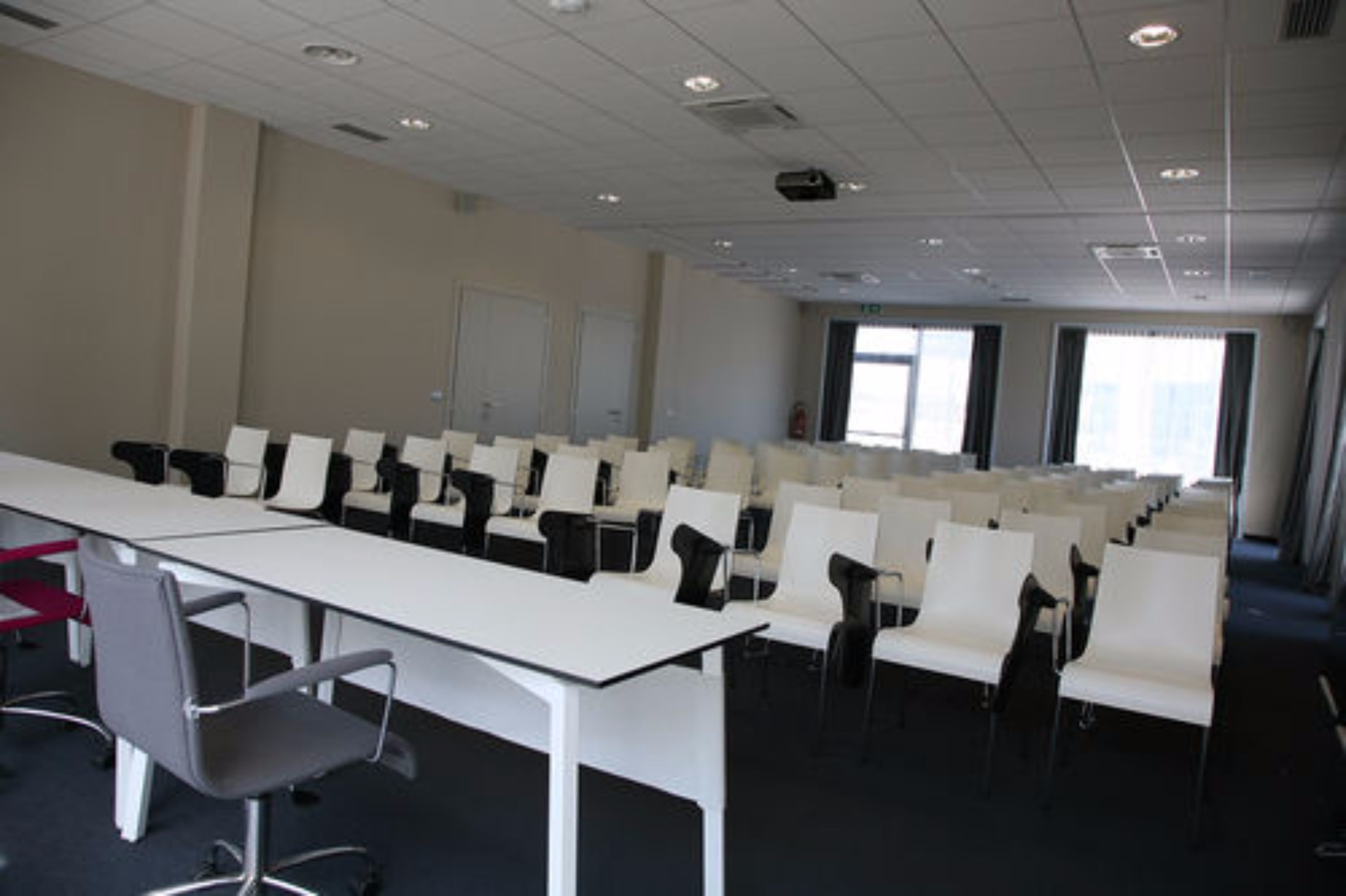 A conference room with rows of white chairs and a long table, large windows, and a door in the background.