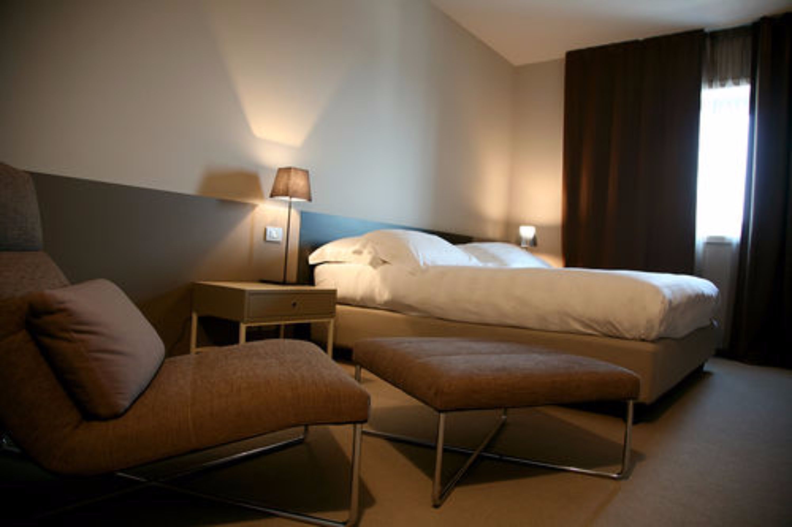 Een hotelkamer met een bed, nachtkastje met lamp, stoel en een voetenbank.