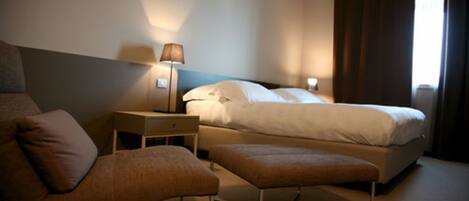 Una habitación de hotel con cama, mesita de noche con lámpara, una silla y un reposapiés.