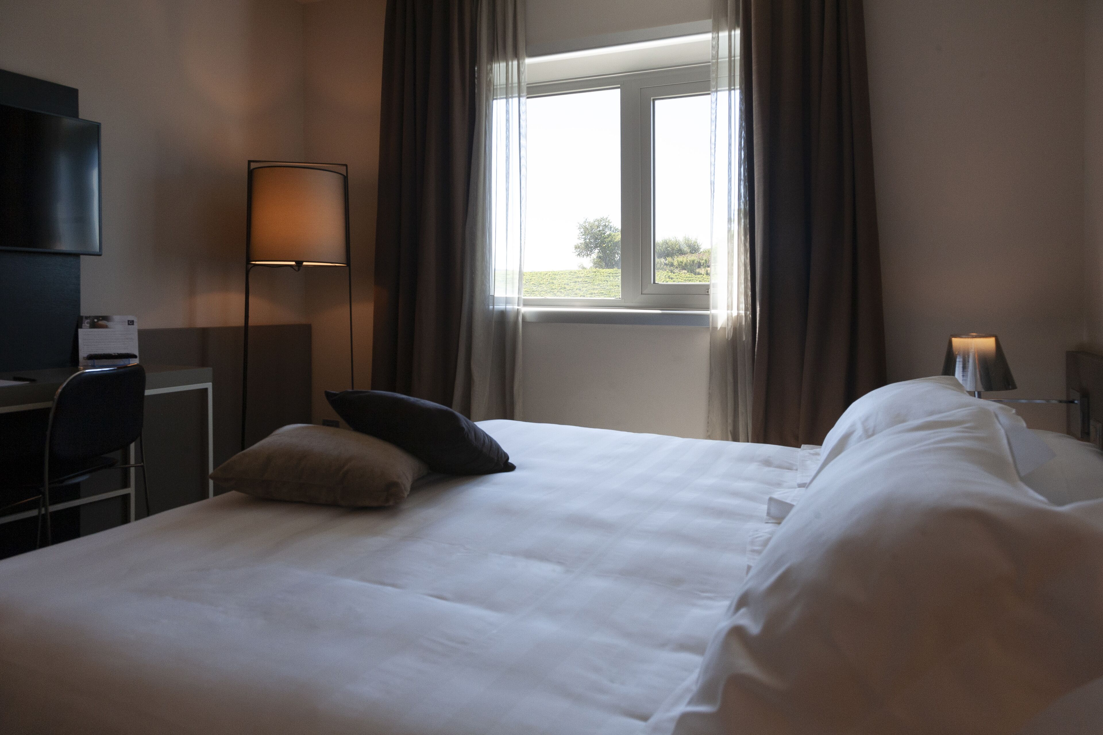 Una habitación de hotel con cama, escritorio, silla, lámpara y ventana con cortinas.