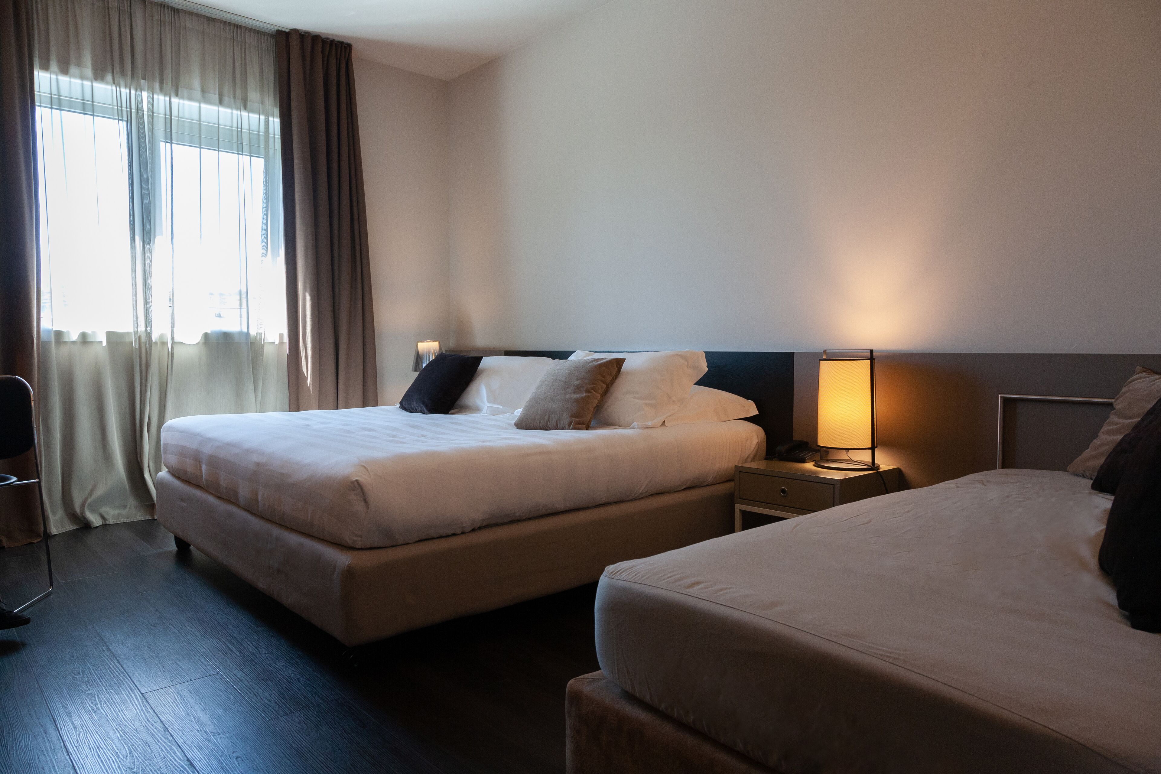 A hotel room with two beds, a bedside table with a lamp, and a window with curtains.
