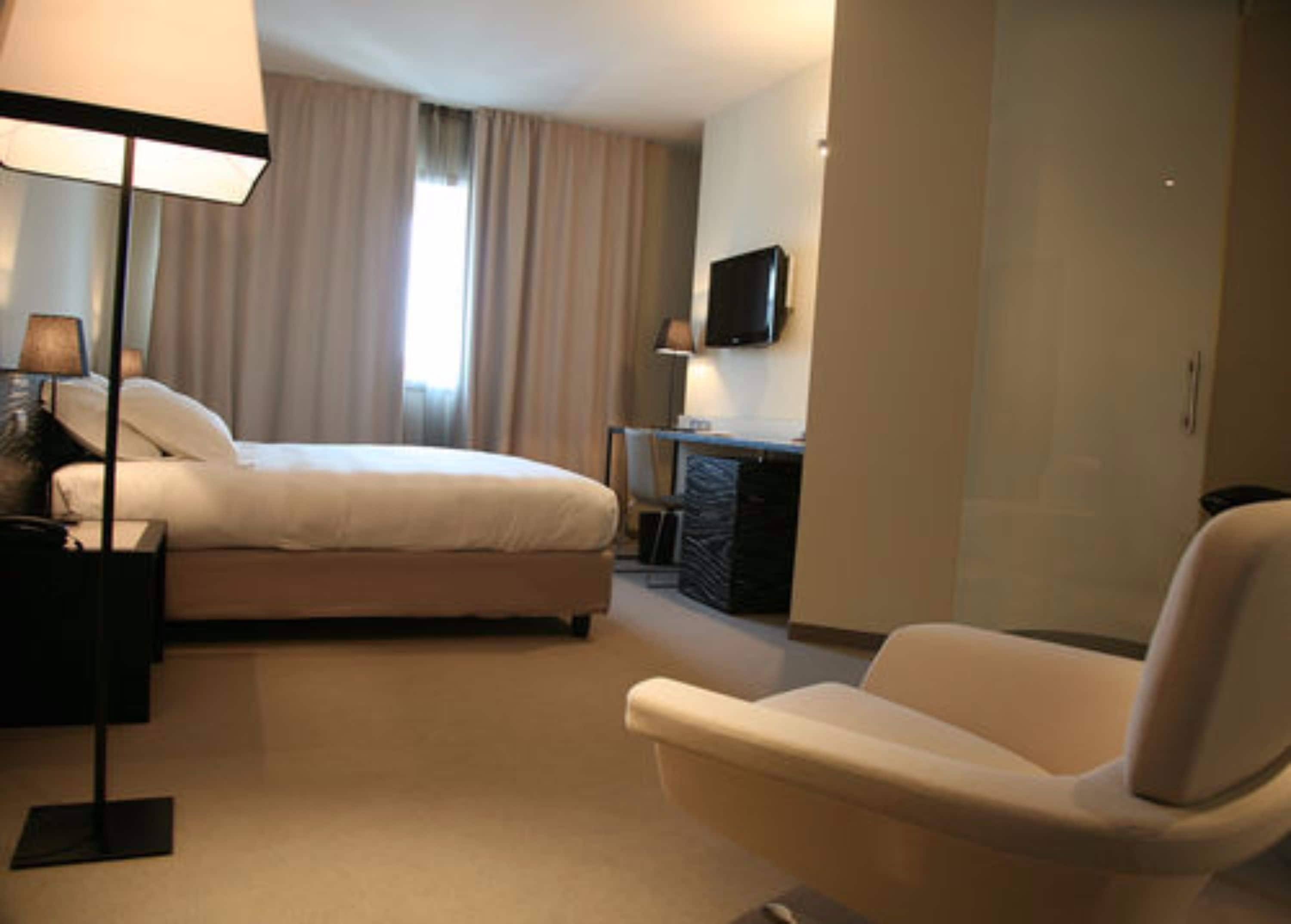A hotel room with a bed, a desk, a chair, a TV, and a lamp.