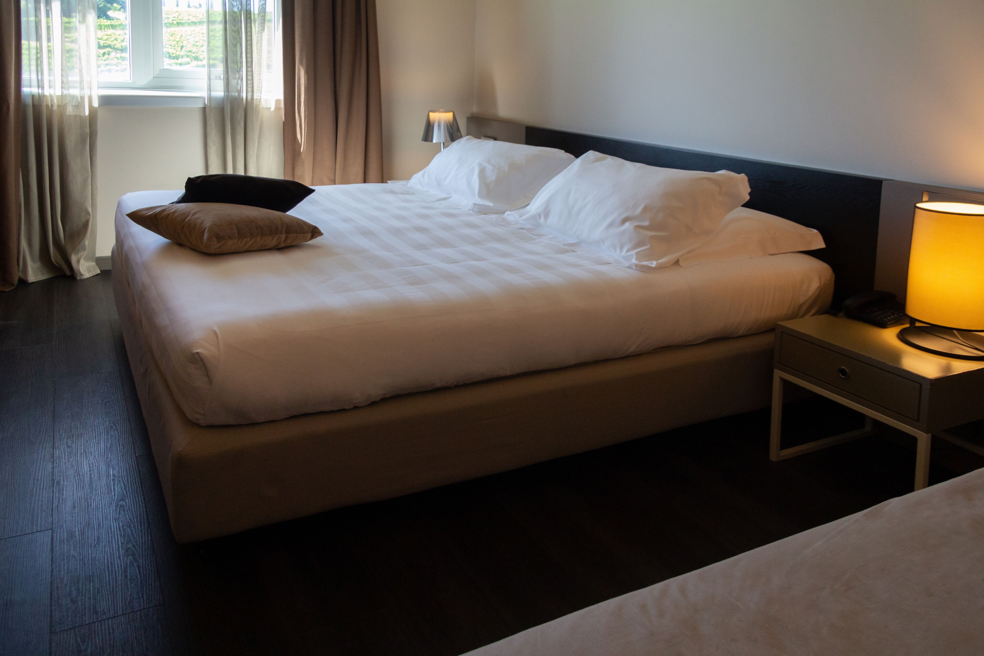 A neatly made bed with white linens and a single brown pillow, a bedside table with a lamp, and a window with curtains.