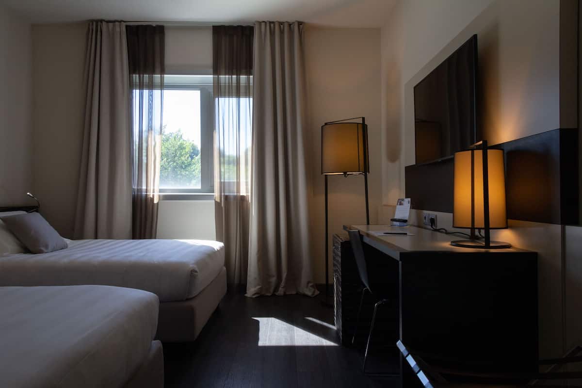 A hotel room with two beds, a desk with a lamp, a flat-screen TV, and a window with curtains.