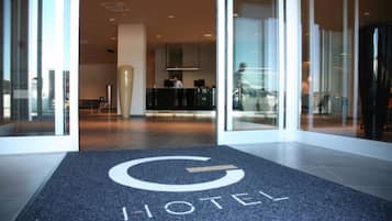 Ein Hotel Eingangsbereich mit Empfangstresen, einer großen Glasdoor und einem Teppich mit dem Hotel-Logo.