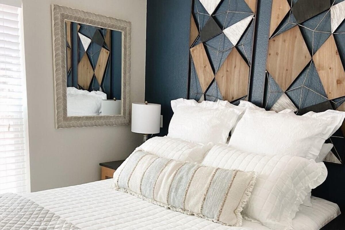 A bedroom with a bed, pillows, a bedside table, a lamp, and a mirror.