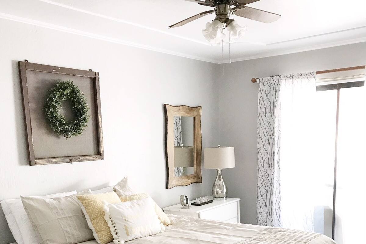 A bedroom with a wooden bed, a ceiling fan, a bedside table with a lamp, a mirror, and a window with curtains.