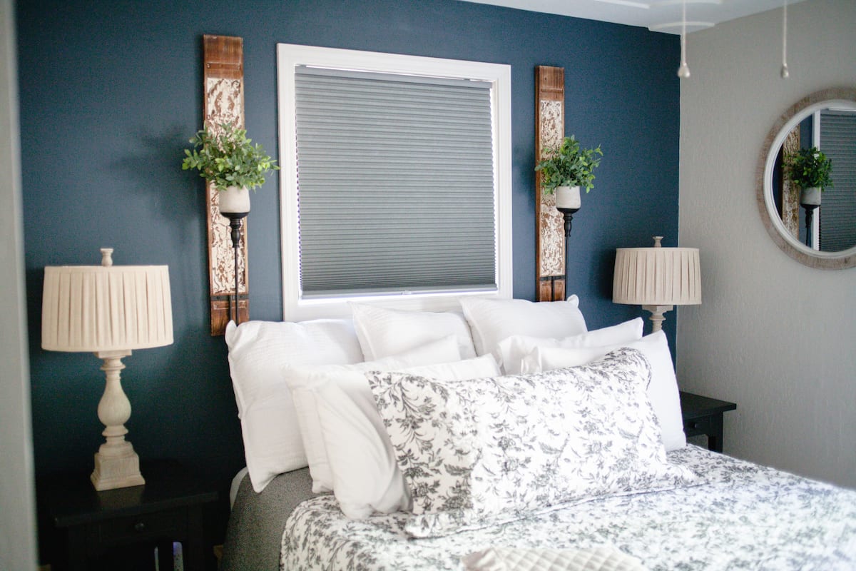 A bedroom with a bed, two bedside lamps, a window with blinds, and two wall-mounted planters.