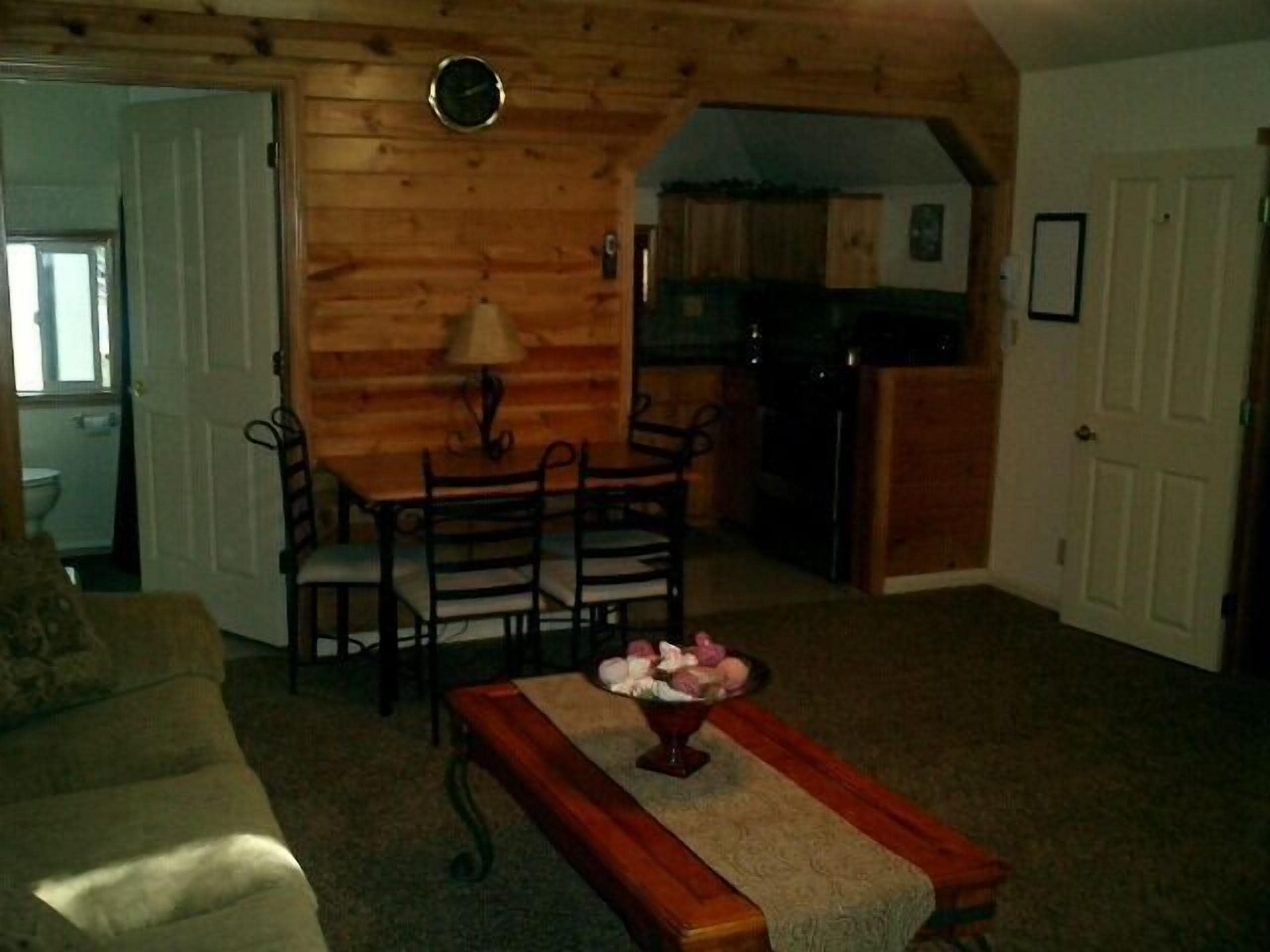 A cozy living room with a wooden ceiling, a dining table with chairs, a coffee table with a vase, and a kitchen in the background.