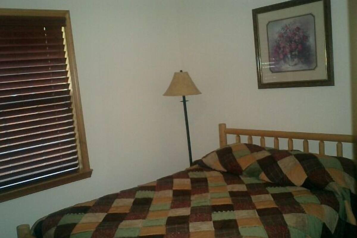 A bedroom with a wooden bed, a checkered bedspread, a lamp, a window with blinds, and a framed picture on the wall.