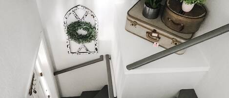 Una escalera con una pared blanca, un adorno de metal en forma de corona y una maleta antigua con plantas encima.