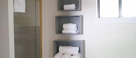 A bathroom with a glass shower door, a toilet, and three shelves with towels.