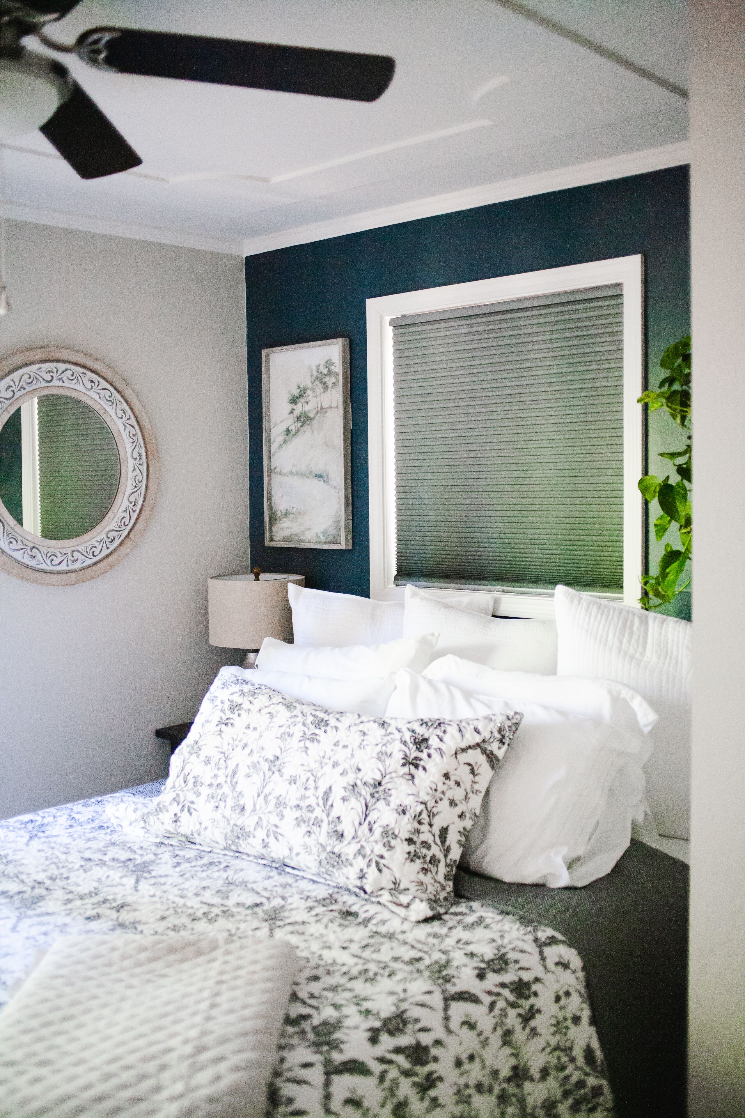 A bedroom with a bed, a round mirror, a window with blinds, a wall-mounted lamp, and a framed picture.