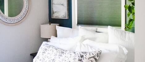 A bedroom with a bed, a round mirror, a window with blinds, a wall-mounted lamp, and a framed picture.