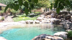 Ein klares Freibad mit Felsen und Wasserfall, umgeben von viel Grün und einem Zaun.
