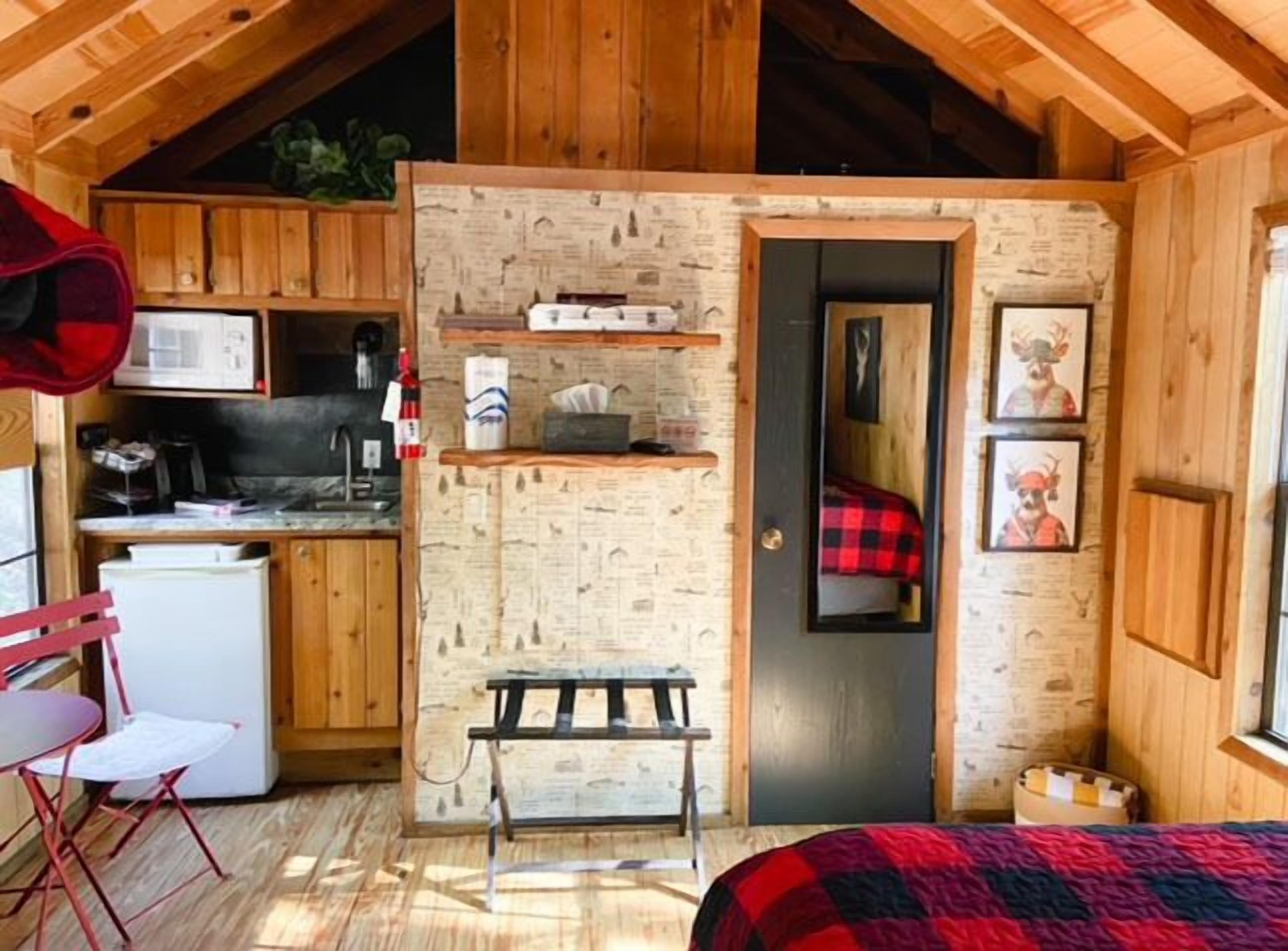 Une petite cabane rustique aux murs en bois et au plafond incliné. Elle comprend un coin cuisine équipé d’un four à micro-ondes, d’un évier et d’un petit réfrigérateur. On y trouve un banc et un lit recouvert d’une couverture à carreaux rouges et noirs.
