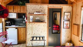 A compact, rustic cabin with wooden walls and a sloped ceiling. It includes a kitchenette with a microwave, sink, and small refrigerator. There is a bench and a bed with a red and black checkered blanket.