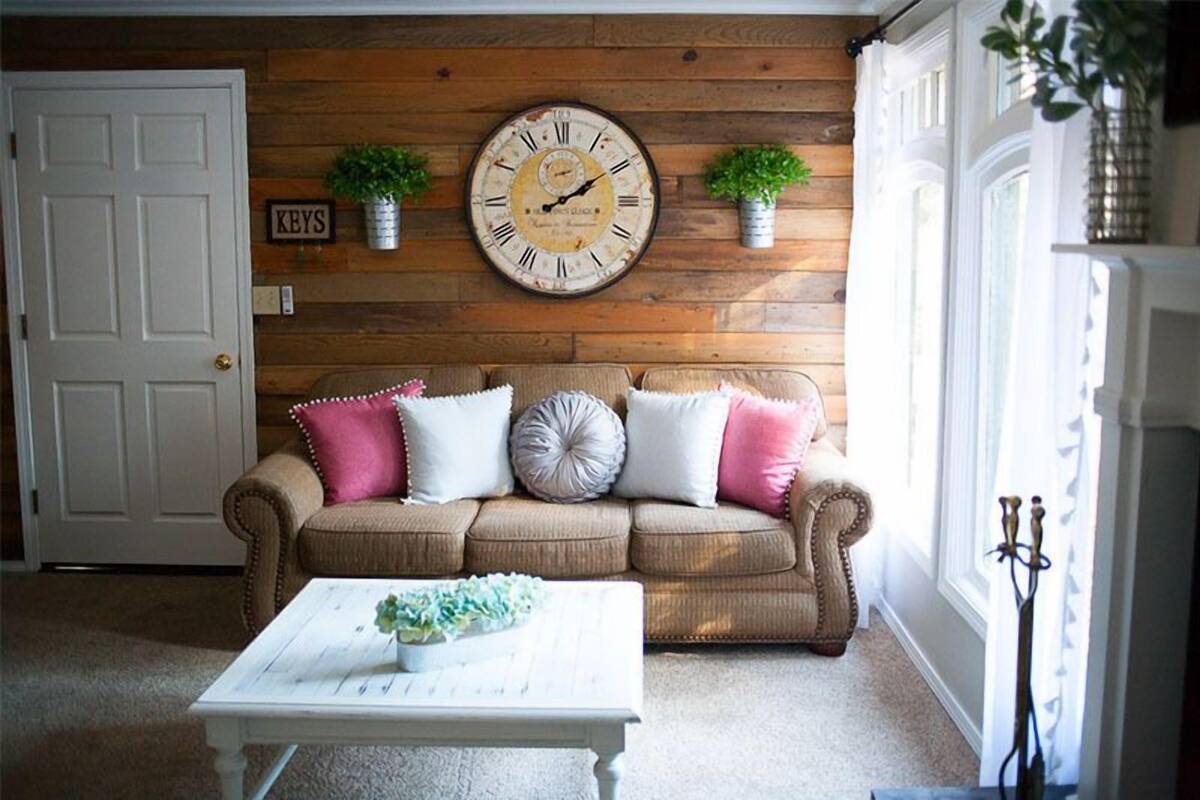 A cozy living room with a wooden wall, a vintage clock, a beige sofa with pink and white cushions, and a white coffee table with a floral arrangement.