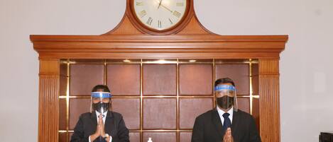Two individuals seated at a desk with a clock above them, a nameplate, and a sign stating no acceptance of American Express cards.