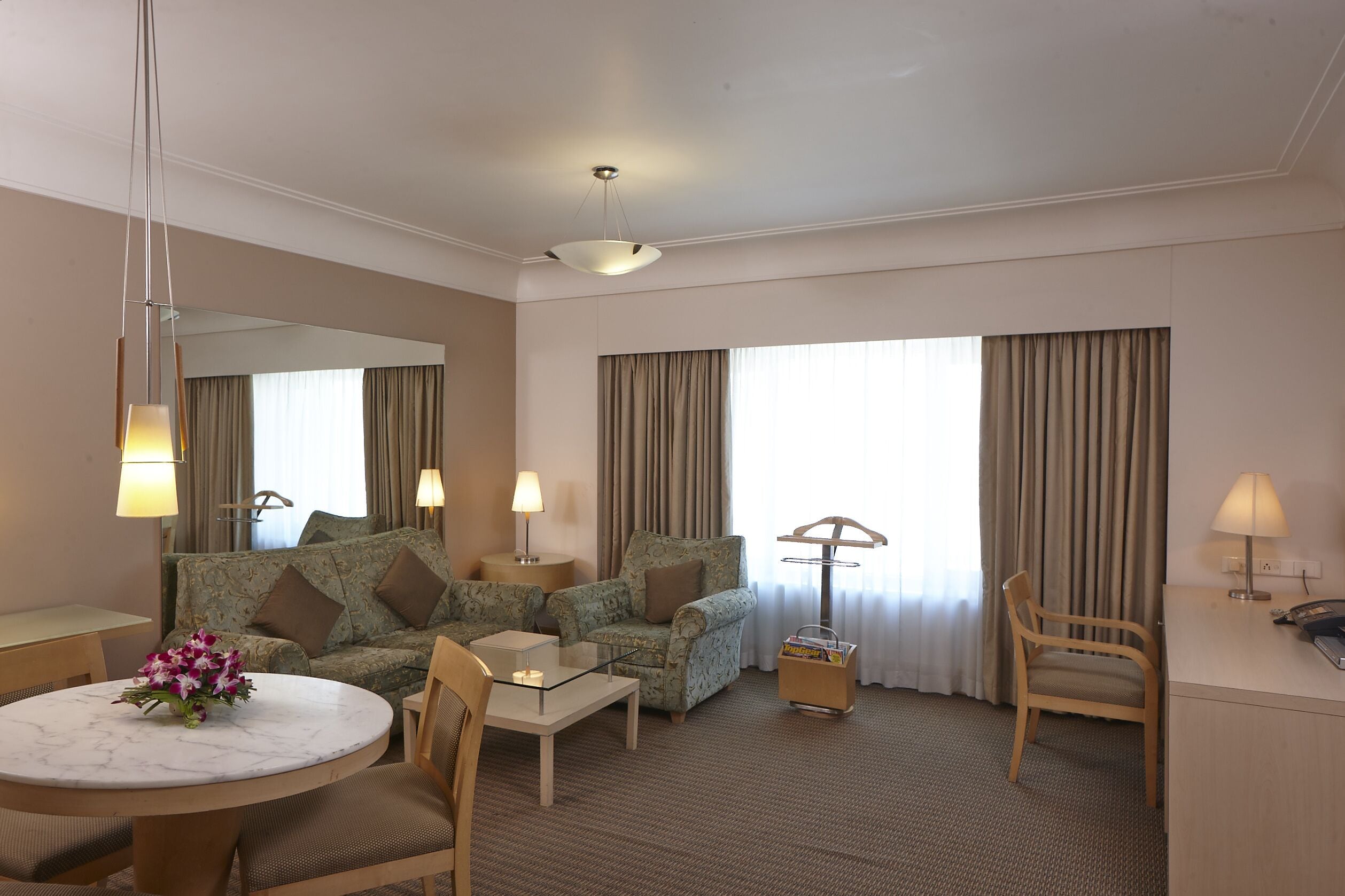 A hotel room with a sofa, armchair, dining table, and a small side table with a lamp.