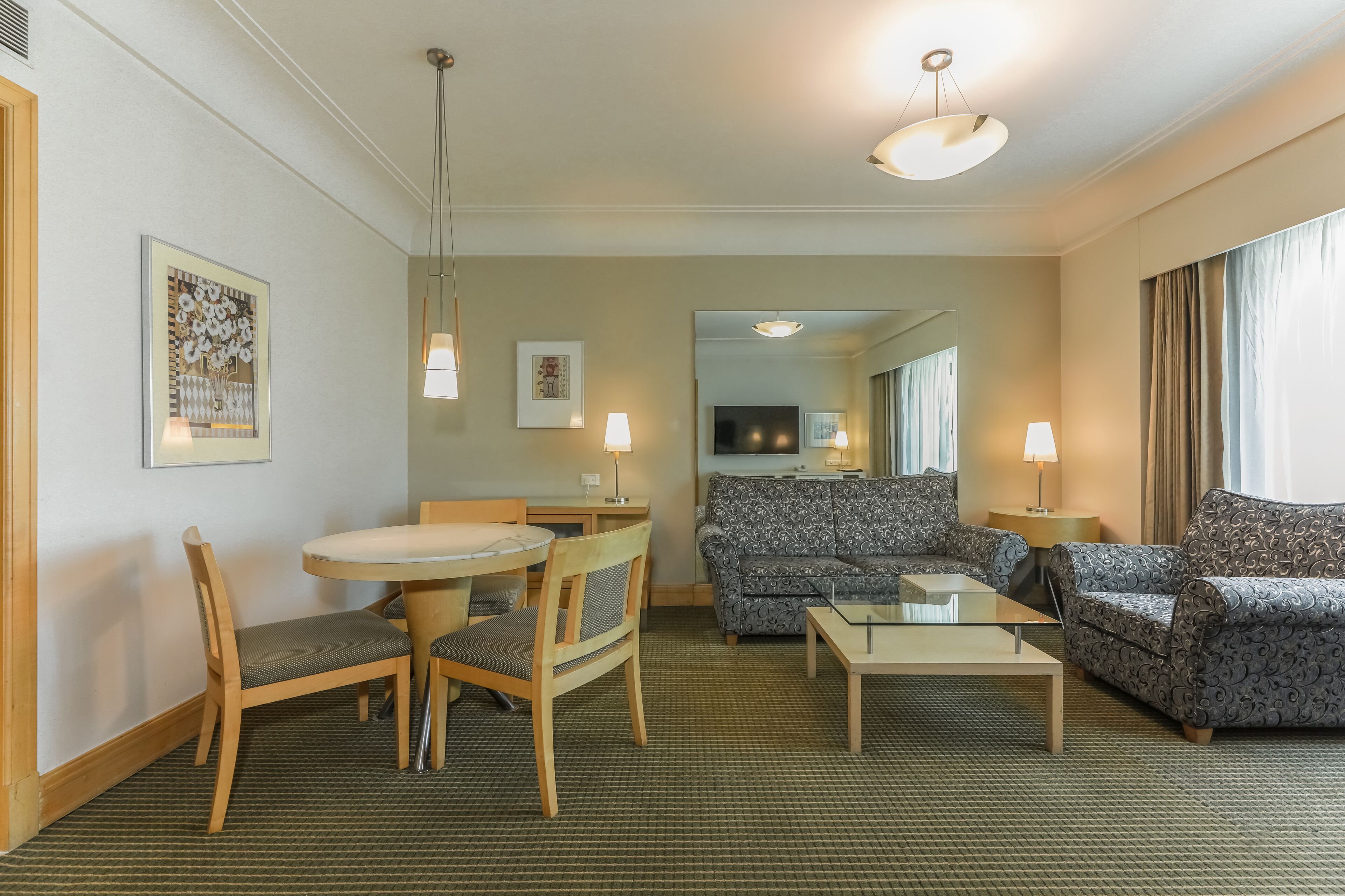 Habitación de hotel con una mesa de comedor, dos sillas, un sofá, una mesa de centro y un televisor al fondo.