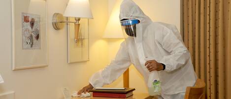 A person in a white protective suit cleaning a wooden table in a room with a lamp and framed artwork on the wall.