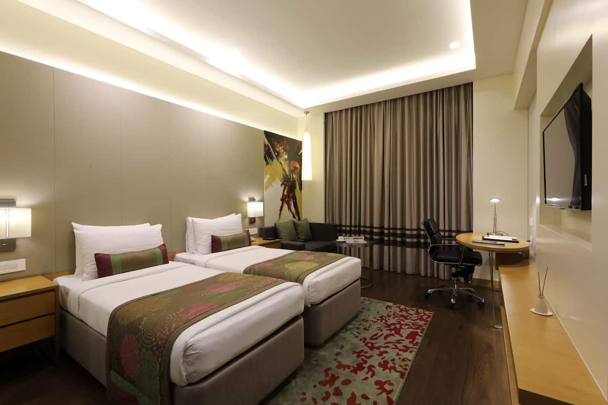 Ein Hotelzimmer mit zwei Betten, einem Schreibtisch, einem Sessel, einem Fernseher und einem großen Fenster mit Vorhängen.