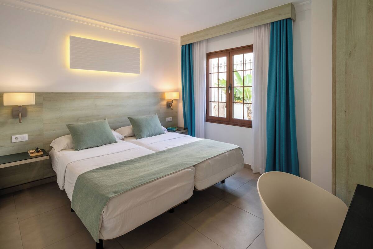Een tweepersoonsbed met een groene deken, twee nachtkastjes met lampen, een raam met blauwe gordijnen en een bad.