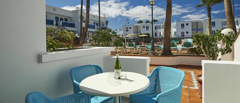 Una terraza con sillas azules y una mesa, con vista a un área de piscina con palmeras y un cielo despejado.