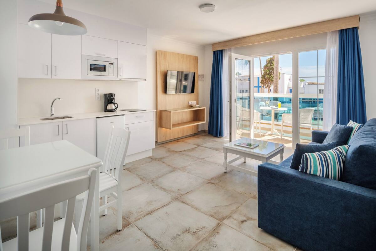 Een moderne hotelkamer met een kitchenette, een eethoek, een zithoek met een bank en een balkon met uitzicht op een zwembad.