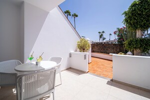 Un patio exterior moderno con paredes blancas, un muro de piedra y suelo de baldosas. Hay dos sillas de mimbre blanco y una pequeña mesa redonda con bebidas.