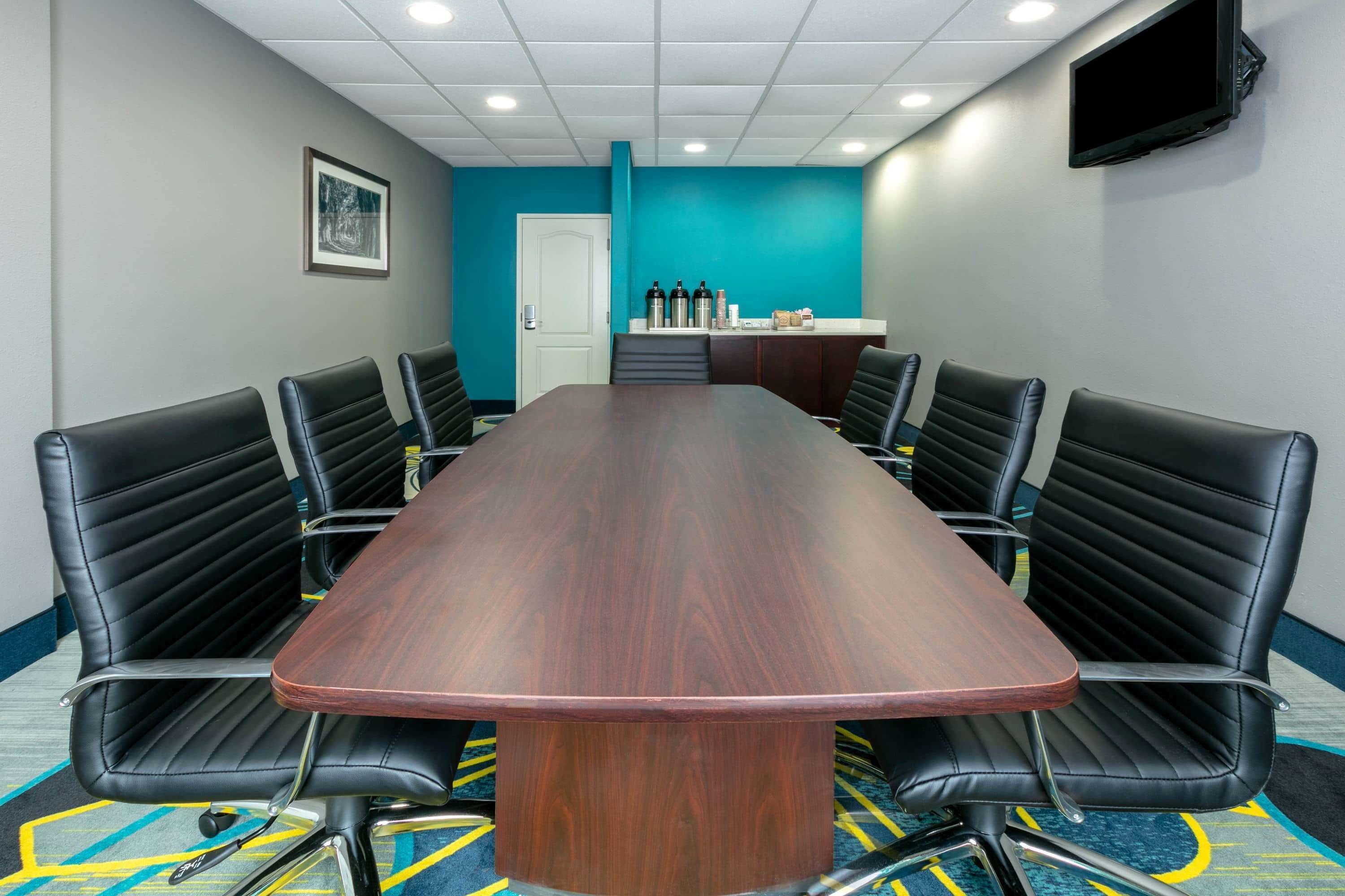Una sala de conferencias con una larga mesa de madera, sillas de cuero negro, una pared de acento color verde azulado, una puerta blanca y un televisor montado.