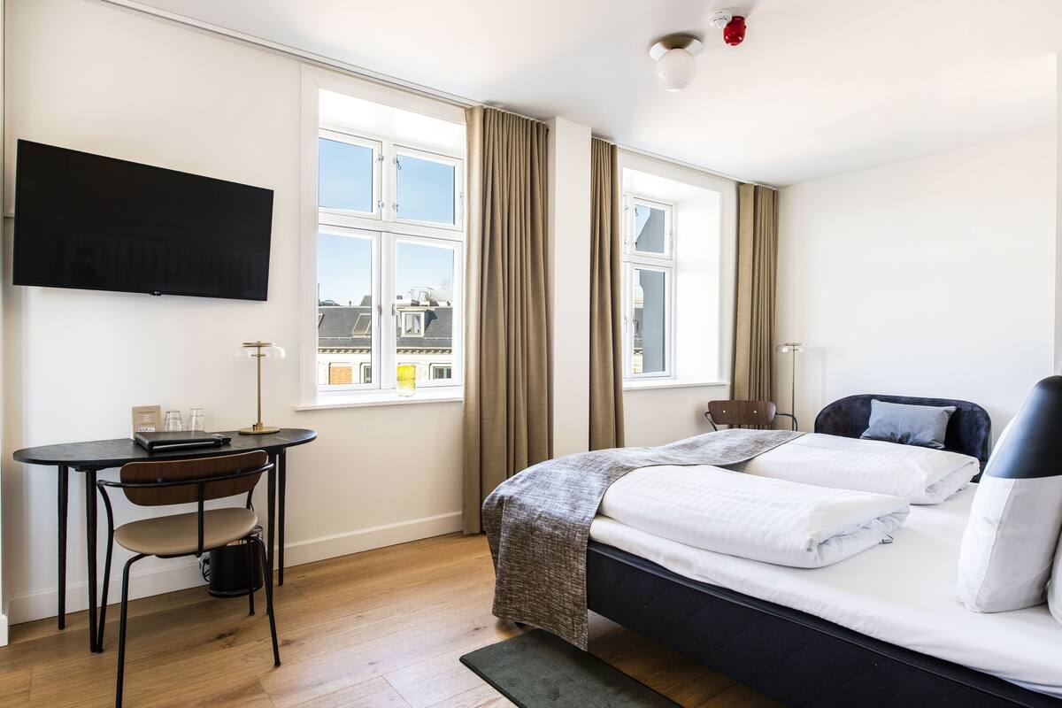 Quarto de hotel com cama, escrivaninha com cadeira, televisão e janelas amplas com cortinas.