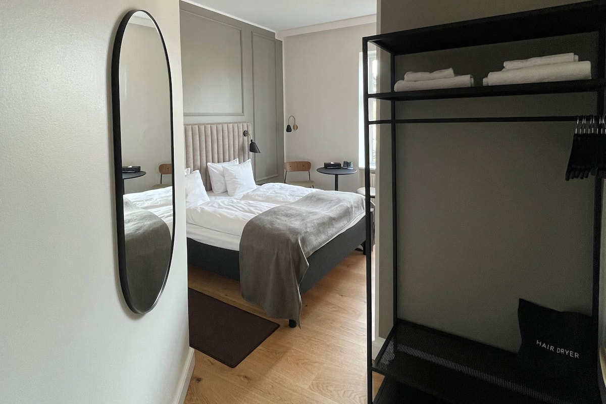 Quarto de hotel moderno com uma cama grande, uma cama beliche, um espelho redondo e uma pequena mesa com abajur.