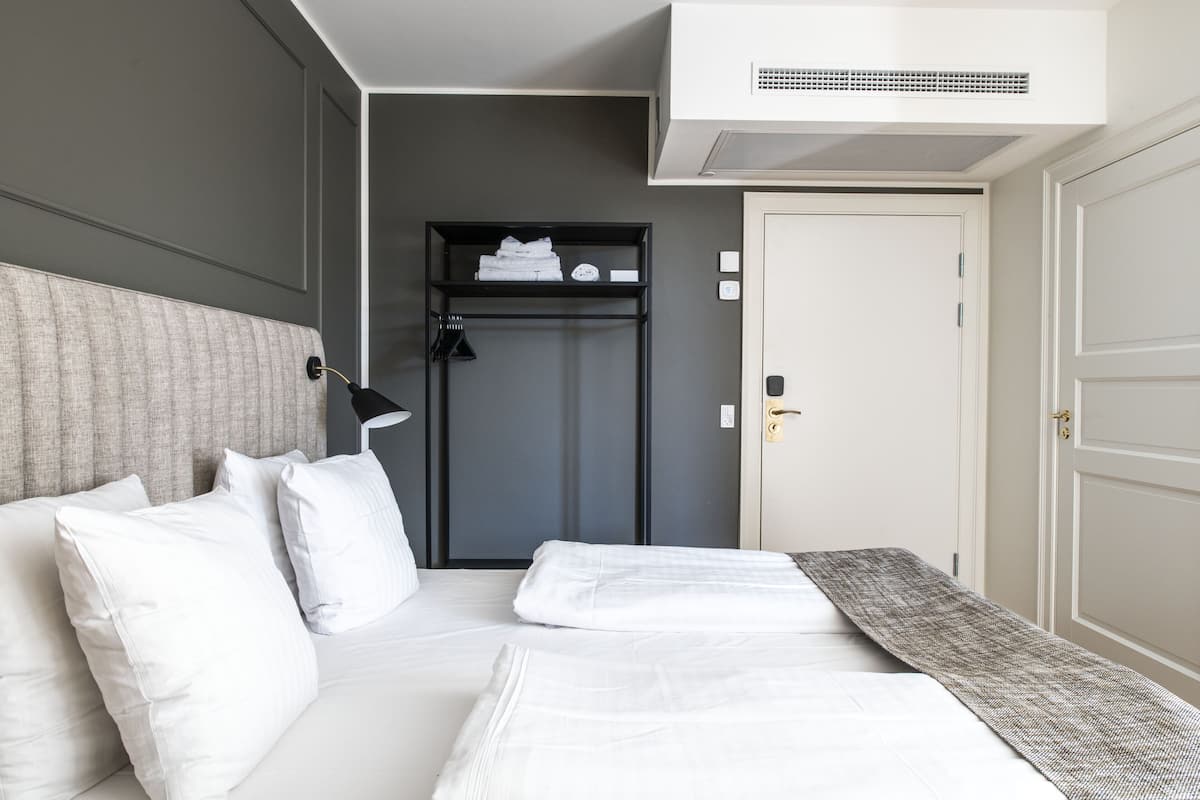 Cama bem arrumada com lençóis brancos, mesa de cabeceira com abajur, prateleira pequena com toalhas dobradas e porta com puxador.