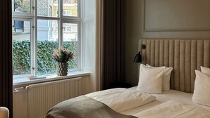 Habitación de hotel con cama, ventana con cortinas, radiador, una mesita con un periódico y una planta en maceta.