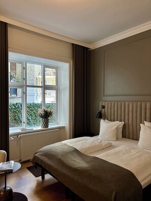Une chambre d’hôtel avec un lit, une fenêtre avec des rideaux, un radiateur, une petite table avec un journal et une plante en pot.