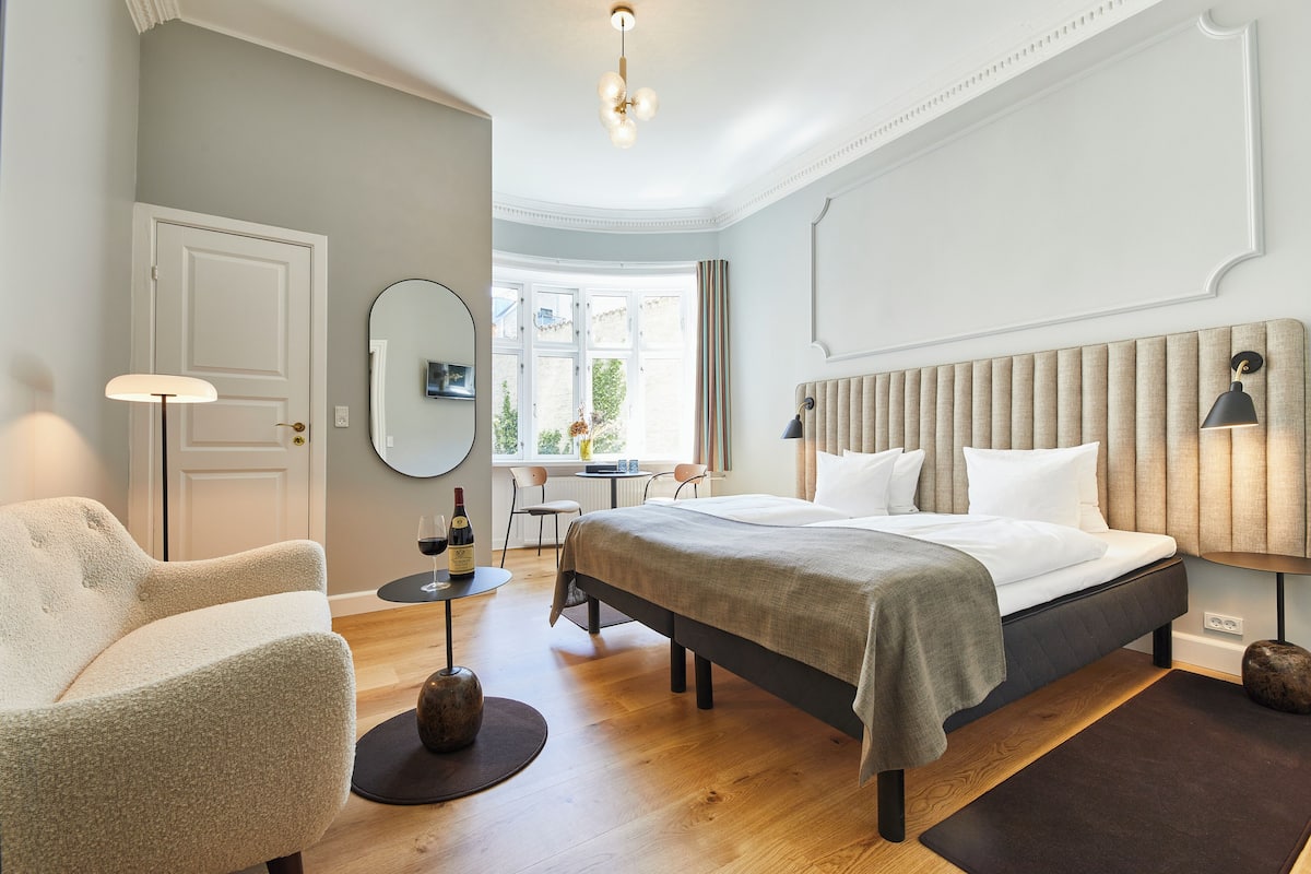 Quarto de hotel com cama, uma cadeira, uma mesa com uma garrafa de vinho e uma janela com cortinas.