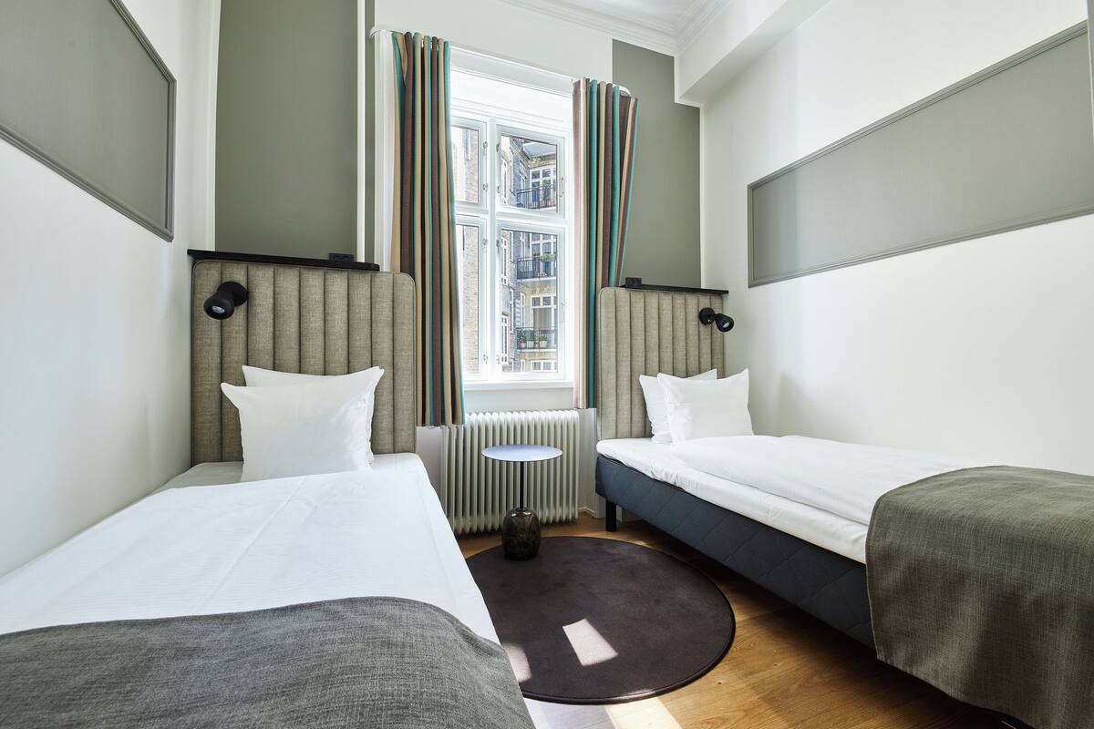 Quarto de hotel com duas camas, uma mesinha redonda, um radiador e uma janela com cortinas.