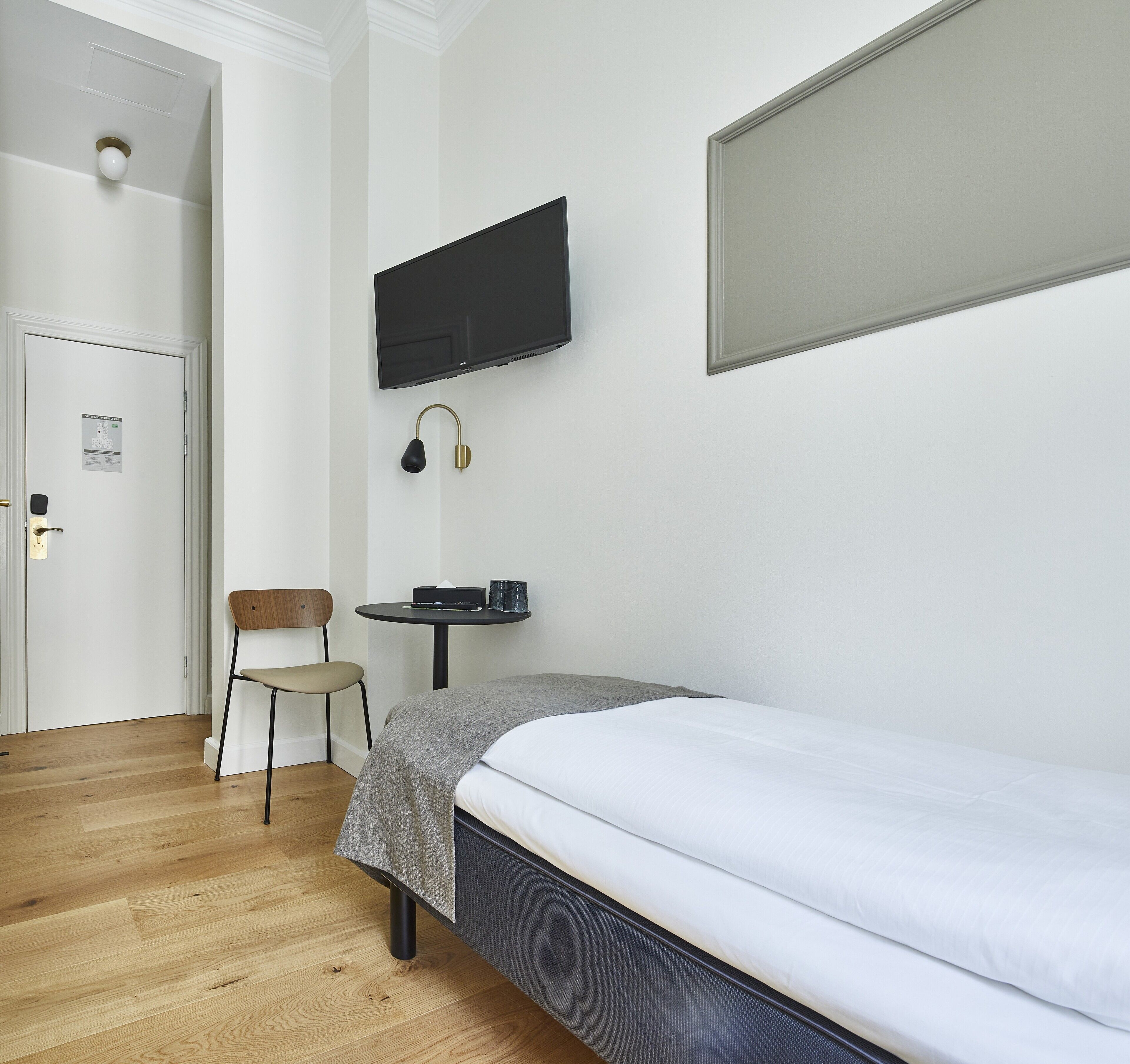 A hotel room with a bed, a desk, a chair, a television mounted on the wall, and a door.
