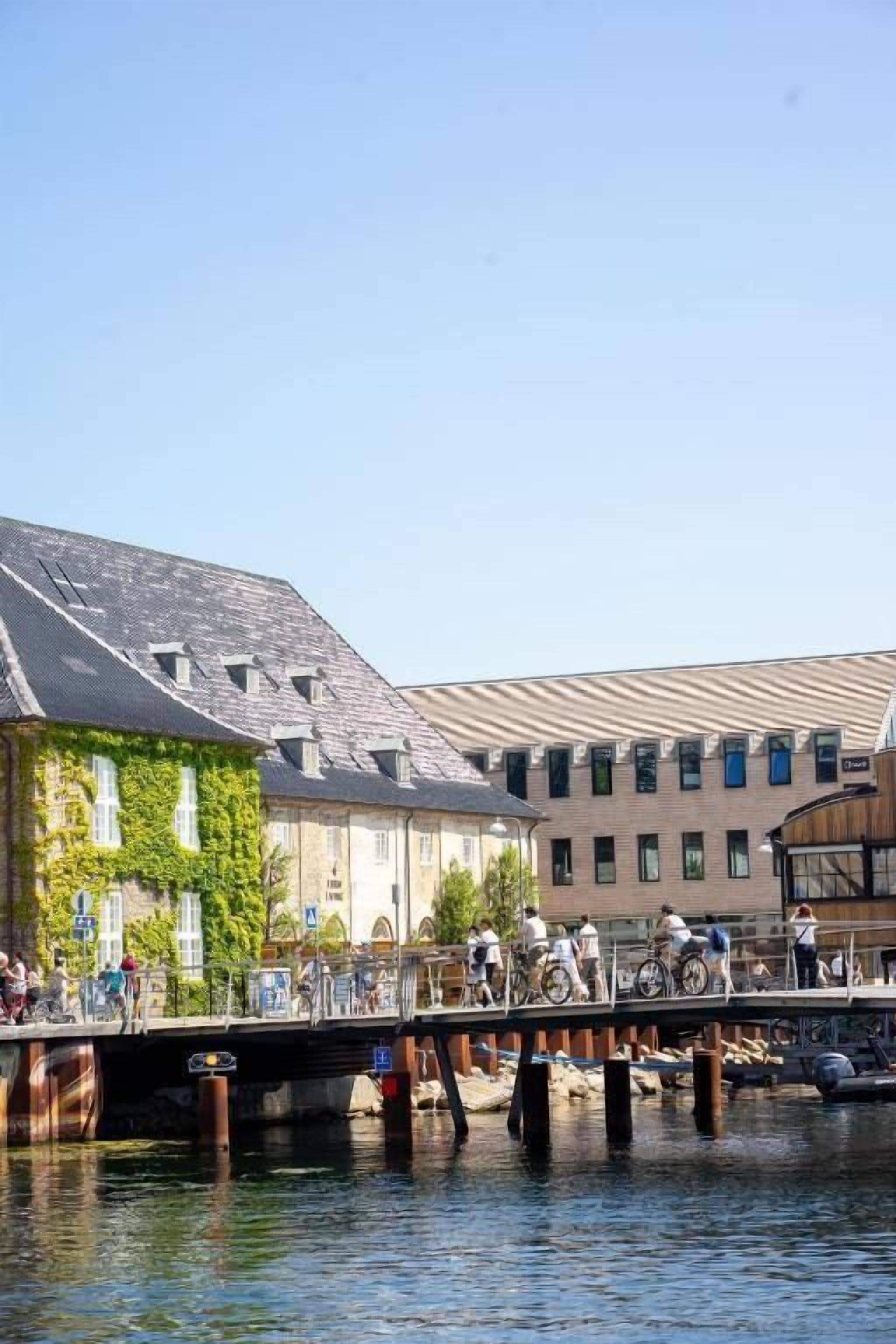 A bridge over water with buildings on either side, a clear sky, and people on bicycles.