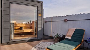 A modern outdoor sauna with a glass door, wooden deck, and a single lounge chair with a blanket. - Hotel Sct Thomas (Frederiksberg)