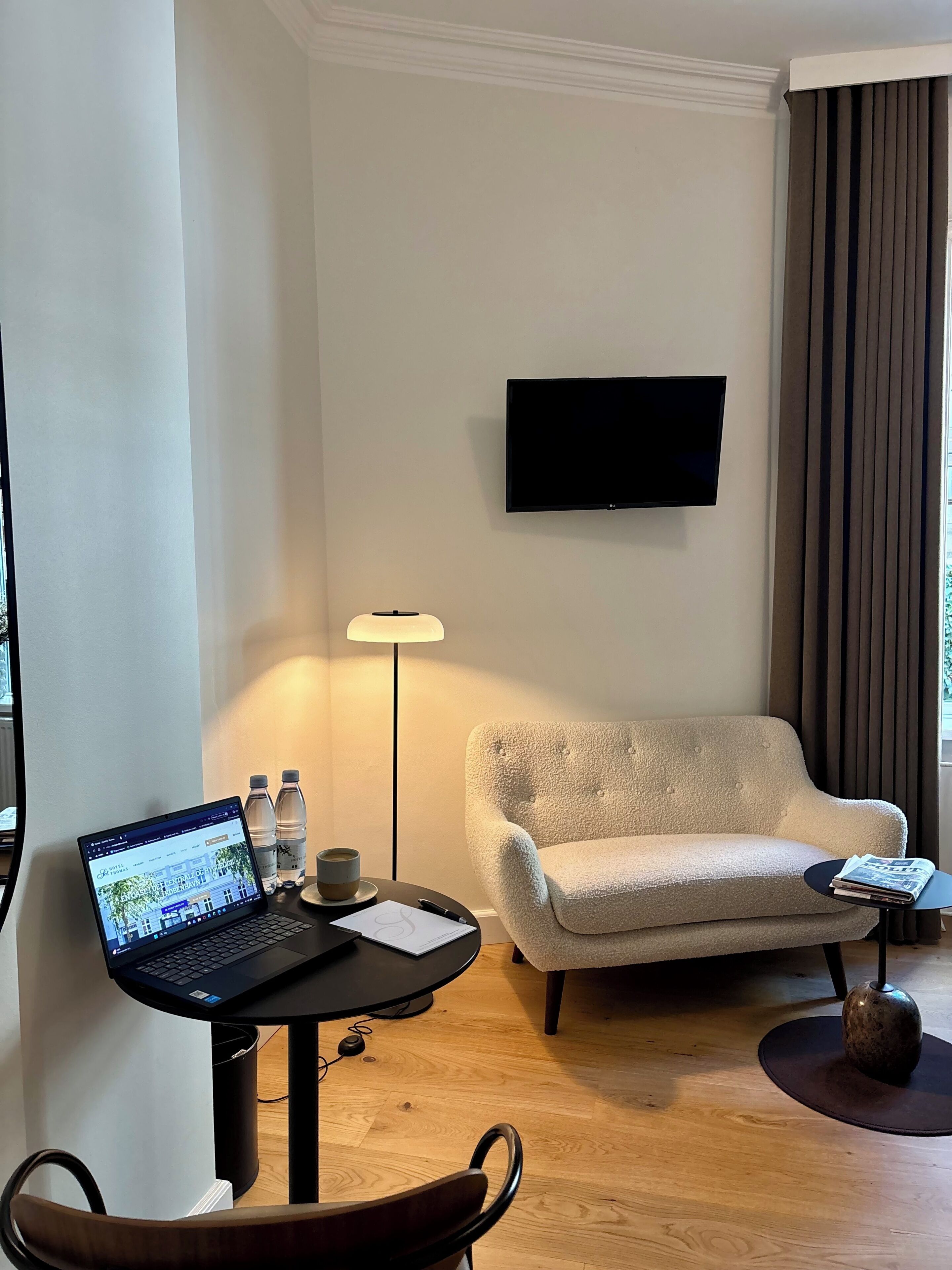 A modern hotel room with a laptop on a desk, a sofa, a TV mounted on the wall, and a lamp.