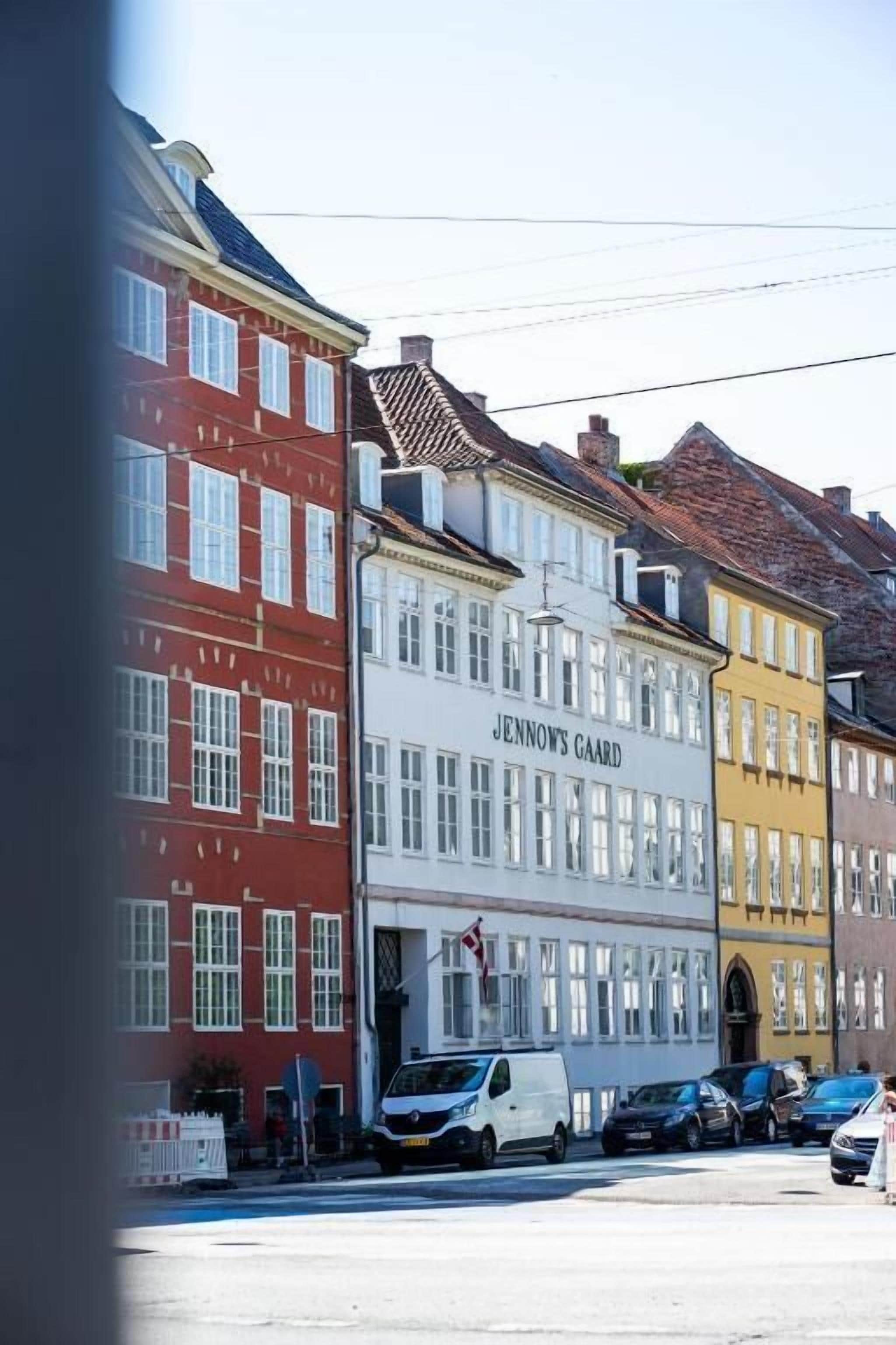 Straßenansicht mit einem Gebäude mit der Aufschrift 'JENNYS GAARD' und geparkten Fahrzeugen.