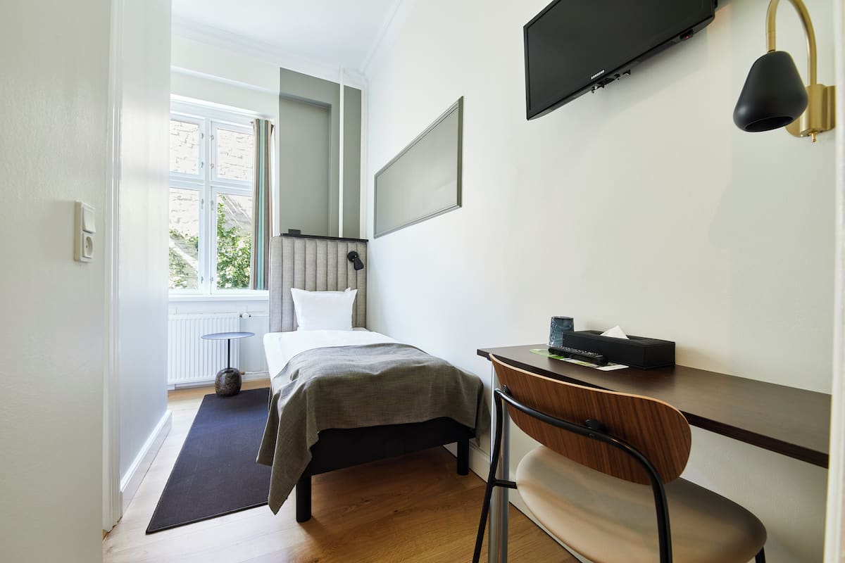 Quarto de hotel com uma cama, uma escrivaninha, uma cadeira, uma televisão e uma janela com vista para árvores.