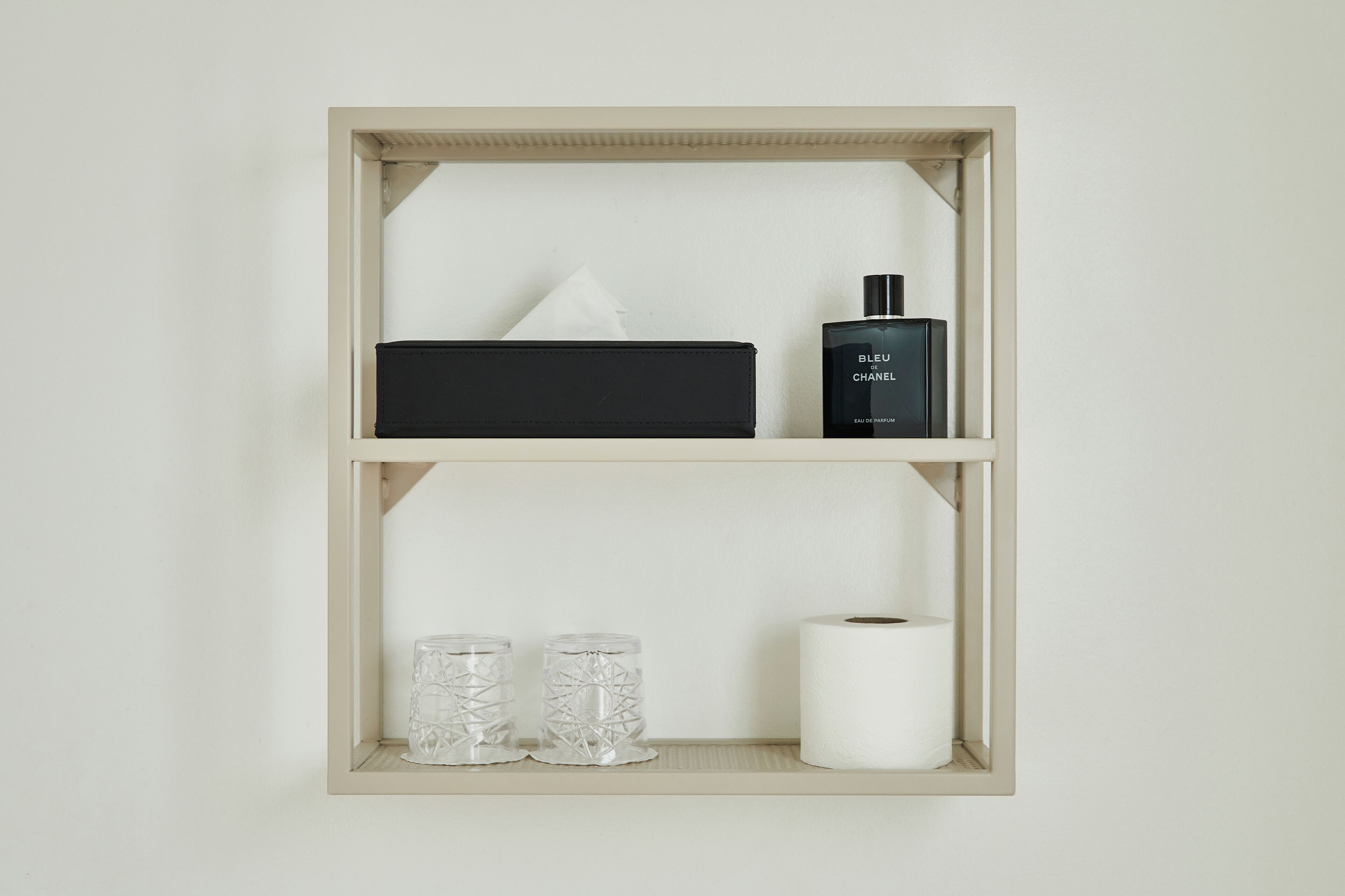 Una estantería con una caja negra, un frasco de perfume Chanel, dos recipientes de vidrio y un rollo de papel higiénico.