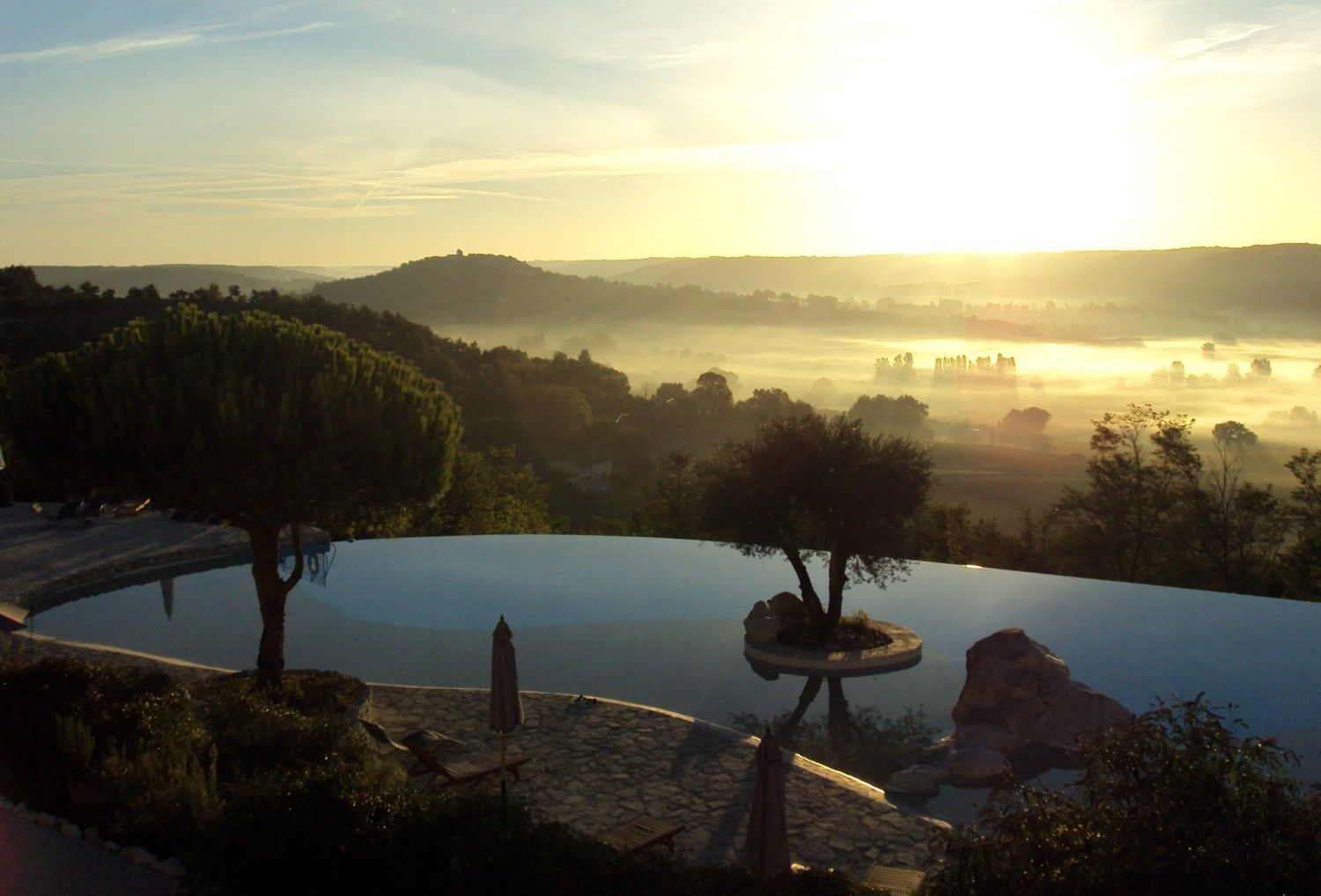 Una piscina circular infinita con borde de piedra, rodeada de árboles y rocas, con vistas a un paisaje brumoso y colinas distantes con un amanecer.
