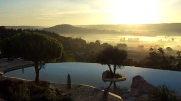 Una piscina circular infinita con borde de piedra, rodeada de árboles y rocas, con vistas a un paisaje brumoso y colinas distantes con un amanecer.