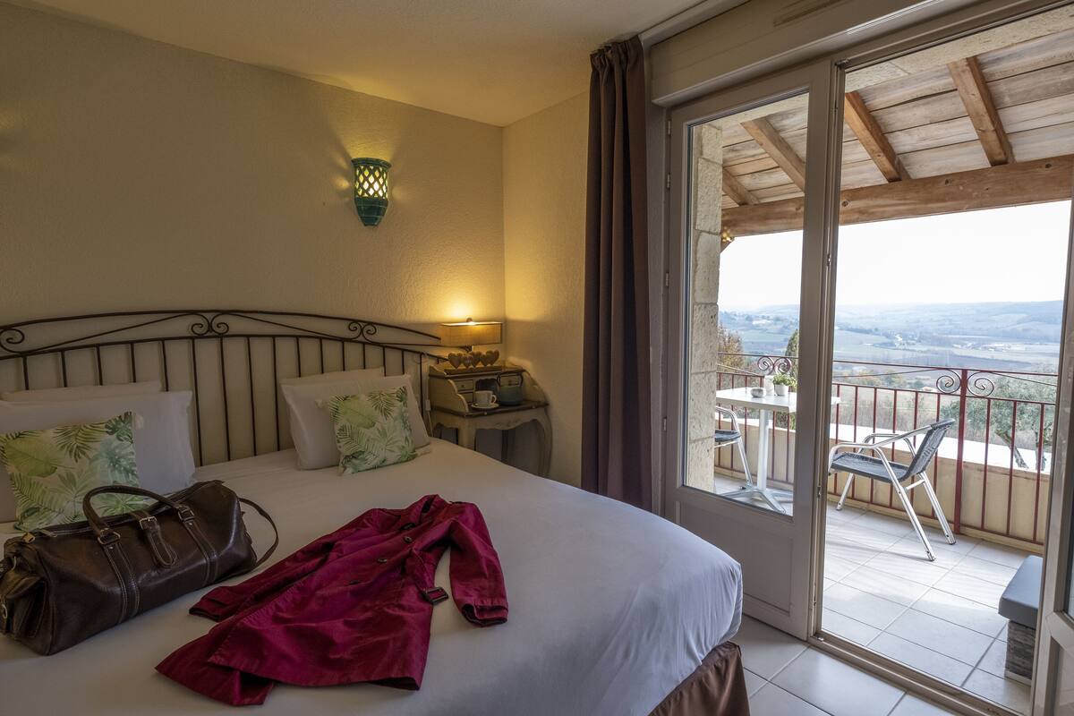 Hotelkamer met een bed, nachtkastje, lamp, uitzicht op de natuur en een balkon met een tafel en stoelen.