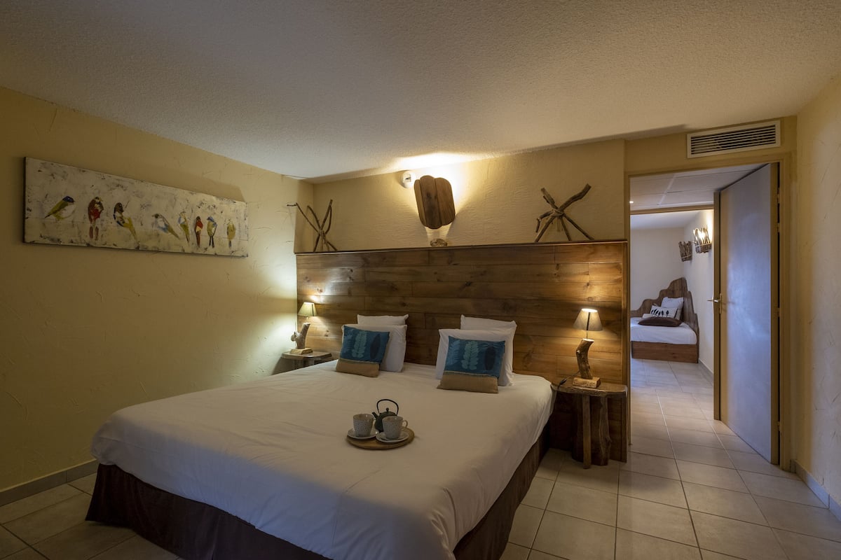 Een hotelkamer met een groot bed, twee nachtkastjes met lampen, een houten hoofdeinde, een schilderij aan de muur en een deur die naar een andere kamer leidt.